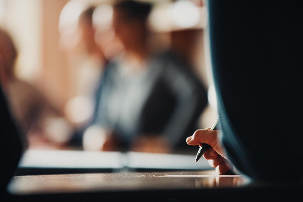 Unscharfer Hintergrund, im Vordergrund jemand, der mit einem Stift in der Hand schreibt.