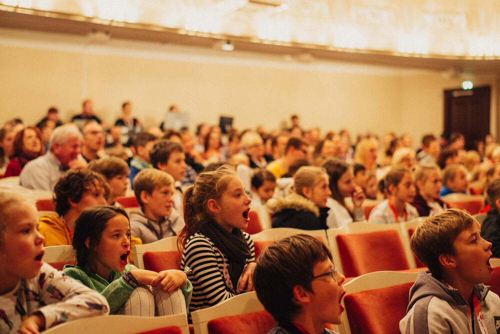 Erwachsene und viele Kinder in einem Theatersaal. Einige Kinder rufen etwas mit weit offenem Mund.